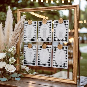 May include: A gold framed seating chart with six cards. Each card has a black and white diamond pattern border and a gold wax seal. The cards display guest names in an oval shape. The frame is on a wooden surface.