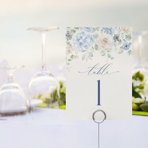 Pode incluir: Um cartão de número de mesa branco com flores de aquarela azul e branca. O cartão diz "mesa 1" em texto azul.