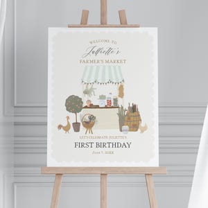 Könnte beinhalten: Dekoratives Schild auf einer Staffelei mit dem Text "Willkommen auf Juliettes Bauernmarkt". Die Illustration zeigt einen Marktstand mit Produkten, einen Baum, Hühnern und den Worten "Erster Geburtstag".