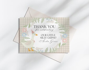 May include: A greeting card with a cartoon goose wearing a party hat, surrounded by a floral wreath. The card reads "THANK YOU for celebrating OUR LITTLE SILLY GOOSE!" in elegant script. The background has a plaid pattern, and the card is placed on an envelope.