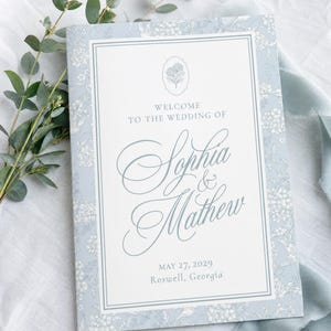 May include: A rectangular wedding programme featuring a light blue floral border and a white centre panel. The text reads: Welcome to the wedding of Sophia & Mathew, May 27, 2029, Roswell, Georgia. A small floral icon is centred at the top.