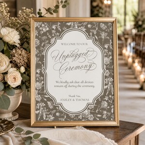 May include: A gold-framed sign with a taupe floral background and a white central panel. The text reads: WELCOME TO OUR Unplugged Ceremony. We kindly ask that all devices remain off during the ceremony. Thank You, ANSLEY & THOMAS.