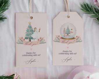 May include: Two pale pink gift tags featuring festive illustrations. One shows a Christmas tree and a ballerina, the other a snow globe. Both have the text "thank you!" and "thanks for celebrating with us!" with a name. They are tied with string.
