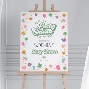 May include: A pink and green baby shower sign with the text "Our Little Lucky Charm is on the way!" and "Welcome to Sophia's Baby Shower" with a date of 05.27.2025. The sign is decorated with a border of green shamrocks, rainbows, hearts, and gold coins.