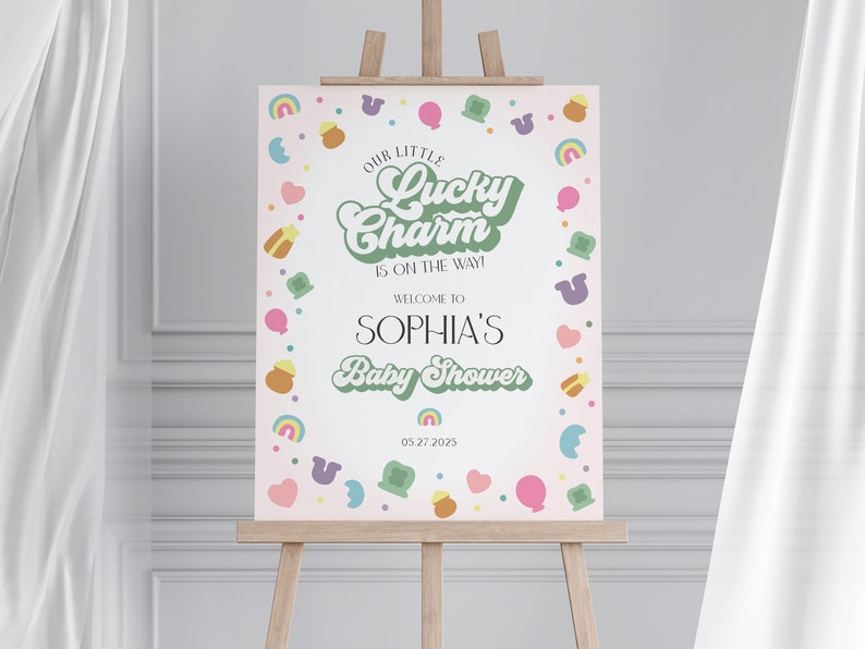 May include: A pink and green baby shower sign with the text "Our Little Lucky Charm is on the way!" and "Welcome to Sophia's Baby Shower" with a date of 05.27.2025. The sign is decorated with a border of green shamrocks, rainbows, hearts, and gold coins.