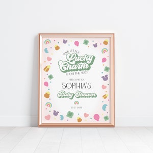 May include: A pink and green baby shower sign with a gold frame. The sign reads "Our Little Lucky Charm is on the way!" and "Welcome to Sophia's Baby Shower" with the date "05272025". The sign is decorated with a border of green shamrocks, rainbows, and gold coins.