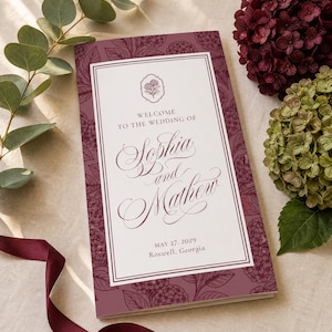 May include: A rectangular wedding program card with a deep burgundy border featuring a white floral pattern. The center white panel displays the text: Welcome to the wedding of Sophia and Mathew, May 27, 2029, Roswell, Georgia, in elegant script.