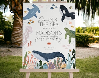 May include: Under the Sea welcome to MADISON'S first birthday party sign with a blue whale, a hammerhead shark, a sea turtle, and a jellyfish. The sign is on a wooden easel.