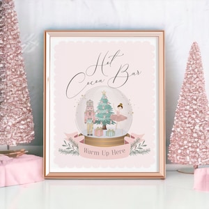 Könnte beinhalten: Ein rosa und goldfarbenes Schild mit dem Text "Hot Cocoa Bar" in Kursivschrift. Das Schild zeigt eine Schneekugel mit einem Nussknacker, einem Weihnachtsbaum und einer Ballerina darin. Der Text "Warm Up Here" befindet sich unter der Schneekugel.