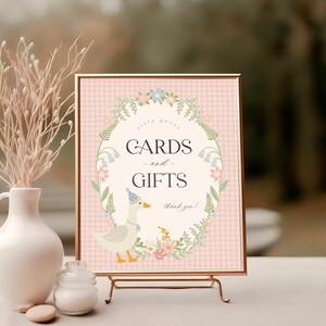 May include: A pink and white gingham sign with a floral border and a cute duck illustration. The sign reads "Cards and Gifts, Thank you!"