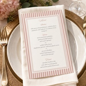 May include: A formal place setting with a white napkin and a menu card. The menu card has a pink striped border and lists starters, entrees, and dessert options. The menu includes baby greens, beef tenderloin, and chocolate hazelnut cake.