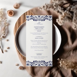 May include: A white menu card with blue floral designs at the top and bottom. The menu card lists the menu for a wedding reception with the names Madelyn & Ryan and the date August 10, 20xx. The menu includes a salad, main course, and dessert.