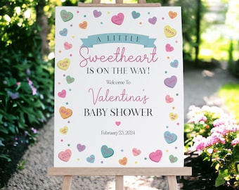 May include: A white sign with pink and blue hearts and the text "A Little Sweetheart Is On The Way! Welcome To Valentina's Baby Shower February 23, 2024".