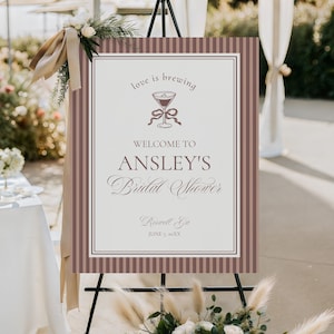 Puede incluir: Un letrero de bienvenida para una despedida de soltera, exhibido en un caballete. El letrero presenta un borde a rayas marrón y blanco y el texto "love is brewing" y "Welcome to Ansley's Bridal Shower". El letrero está decorado con detalles florales.