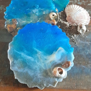 Puede incluir: Cuatro posavasos de resina azul con remolinos blancos y marrones, que se asemejan a las olas del océano, con conchas incrustadas en la resina.