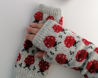 May include: Gray knitted fingerless gloves with a repeating pattern of red ladybugs with black details. The gloves are made of a soft, textured yarn. A small, gray stone is in the background.