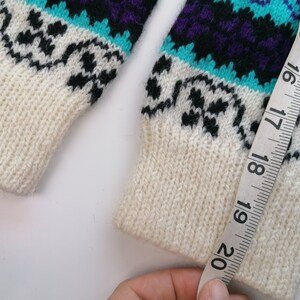 May include: White knitted sweater with a black, purple, and turquoise geometric pattern. A measuring tape is wrapped around the sleeve, showing a measurement of 20 inches.