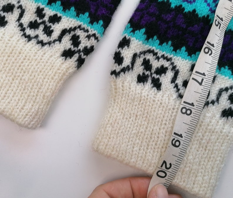 May include: White knitted sweater with a black, purple, and turquoise geometric pattern. A measuring tape is wrapped around the sleeve, showing a measurement of 20 inches.