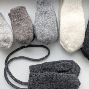 Könnte beinhalten: Eine Sammlung gestrickter Fäustlinge in verschiedenen Grau- und Off-White-Tönen. Ein Paar dunkelgraue Fäustlinge ist durch eine Strickkordel verbunden. Die Fäustlinge sind auf einer weißen Oberfläche angeordnet.