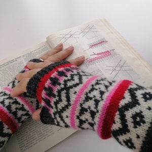 May include: A pair of black, white, pink and red fingerless gloves with a geometric pattern. The gloves are being held by a person who is reading a book with knitting instructions.