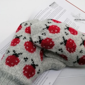 May include: A pair of gray knitted mittens with a red ladybug pattern. The mittens are lying on a white surface with text in a foreign language.