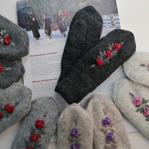 May include: Five pairs of knitted mittens with floral embroidery. The mittens are in various shades of grey and beige. Some mittens have red flowers, while others have purple flowers.
