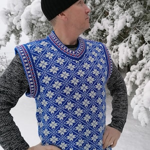 Puede incluir: Un chaleco sin mangas azul y blanco con estampado y ribete rojo y blanco. El chaleco lo lleva puesto una persona que lleva una boina negra y unos vaqueros azules. La persona está de pie frente a un pino nevado.