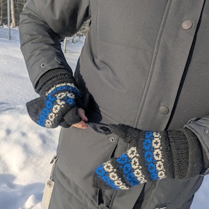 May include: A pair of black knitted mittens with a blue and white geometric pattern. The mittens are worn by a person wearing a dark gray winter coat. The mittens are designed to keep hands warm in cold weather.