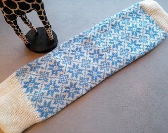 May include: A white and blue knitted leg warmer with a snowflake pattern. The leg warmer is folded in half and lying on a gray surface.