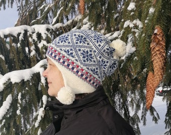 May include: A white and blue knitted hat with a geometric pattern and red accents. The hat has ear flaps with white pom-poms.
