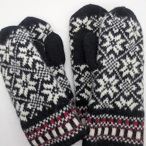 Könnte beinhalten: Ein Paar schwarz-weiße Strickfäustlinge mit Schneeflockenmuster. Die Fäustlinge haben schwarze Daumen und Bündchen sowie ein rot-weiß gemustertes Band am Handgelenk. Diese Winteraccessoires sind ideal für kaltes Wetter.