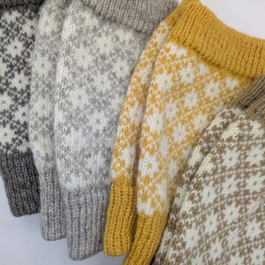 Könnte beinhalten: Nahaufnahme von vier gestrickten Fäustlingen in verschiedenen Farben. Die Fäustlinge weisen ein Rautenmuster in Weiß und Grau, Weiß und Gelb sowie Weiß und Braun auf. Jeder Fäustling hat einfarbige Bündchen.