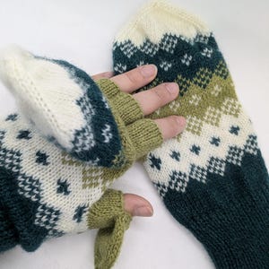 May include: A pair of knitted fingerless gloves with a geometric pattern. The gloves feature a color scheme of white, dark teal, and olive green. The design includes diamond and star-like shapes. One glove is laid flat, while the other is being worn.