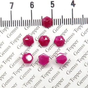 May include: Seven faceted ruby red gemstones, each approximately 5mm in diameter, are arranged on a white surface with a ruler for scale. The ruler is marked with numbers 4, 5, 6, and 7.