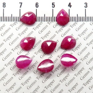 Pode incluir: Um grupo de dez pedras preciosas de rubi em forma de pêra, variando de 6 mm a 8 mm, são exibidas em uma superfície branca com uma régua para escala. As pedras preciosas são de cor vermelha profunda e têm um corte facetado.
