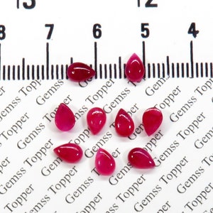 May include: Nine pink teardrop-shaped gemstones on a white surface with a ruler marked in inches.