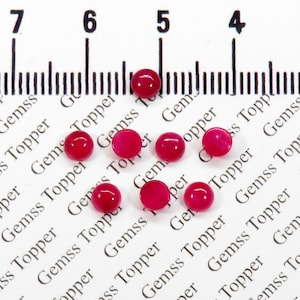 May include: Eight small, round, red gemstones on a white surface with a ruler marked in inches. The gemstones are approximately 1/4 inch in diameter.