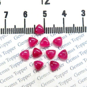 Op de afbeelding: Een groep van 12 roze driehoekige edelstenen, elk ongeveer 5 mm groot, zijn gerangschikt op een wit oppervlak met een liniaal op de achtergrond.