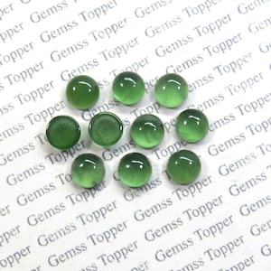 Puede incluir: Diez piedras preciosas cabujón redondas de color verde, cada una con un tono de verde ligeramente diferente. Las piedras preciosas están dispersas sobre una superficie blanca con el texto "Gemss Topper" repetido.