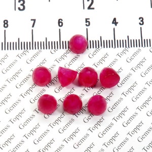 May include: A group of nine pink cabochon gemstones, some are round and some are half-round, on a white surface with a ruler in the background.
