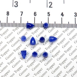 Op de afbeelding: Een verzameling van tien blauwe edelsteen cabochons in verschillende vormen en maten, waaronder druppelvormig, rond en ovaal, gerangschikt op een wit oppervlak met een liniaal voor schaal.