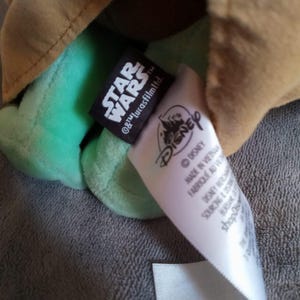 May include: A green plush toy character with a tan robe. The toy has a black tag with "STAR WARS" and "&copy; & &trade; Lucasfilm Ltd." printed on it. A white tag with "DISNEY" is also visible. The toy is made in Vietnam.