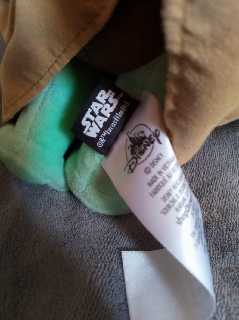 May include: A green plush toy character with a tan robe. The toy has a black tag with "STAR WARS" and "&copy; & &trade; Lucasfilm Ltd." printed on it. A white tag with "DISNEY" is also visible. The toy is made in Vietnam.
