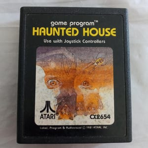 Może przedstawiać: Czarny wkład do gry Atari z żółtą etykietą, na której widnieje napis "game program™ HAUNTED HOUSE Use with Joystick Controllers ATARI CX2654 Label, Program & Audiovisual © 1981 ATARI, INC.". Etykieta zawiera kolorową ilustrację twarzy z oczami i nietoperzem.
