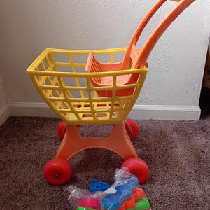 May include: A bright yellow and orange plastic toy shopping cart with red wheels. The cart has a basket, a seat, and a handle. A bag of colorful plastic toy food is in front of the cart.