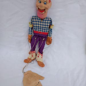 May include: A vintage marionette puppet with a painted face, red hair, and a checkered shirt. The puppet wears purple pants, brown boots, and a red belt. A detached hand-shaped control is visible below.