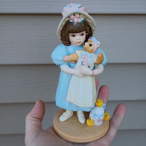 Könnte beinhalten: Porzellanfigur eines Mädchens in hellblauem Kleid und weißem Hut mit rosa Blumen, das einen Teddybären hält. Die Figur steht auf einem Holzsockel mit einem kleinen Krug und drei gelben Küken. Die Figur ist etwa 13 cm hoch.