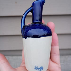 Puede incluir: Una pequeña jarra de cerámica con la parte superior y el asa de color azul cobalto y una base blanca. La jarra tiene un pico curvo y un asa pequeña. La palabra "Doffs" está escrita en escritura azul en la base.