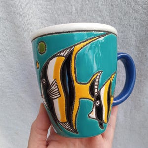 May include: A ceramic mug with a teal background and a blue handle. The mug features a colorful design of two fish with yellow, black, and white stripes. The rim is white with a black trim.