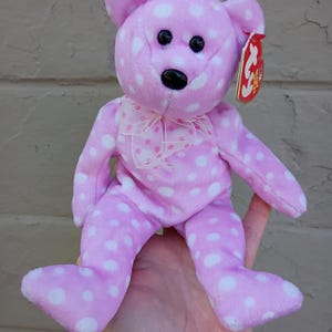 Könnte beinhalten: Ein rosa Teddybär mit weißen Punkten. Der Bär hat schwarze Augen und eine schwarze Nase. Er hat ein rosa Band um den Hals und ein rotes Etikett am Ohr. Ein Stofftier.